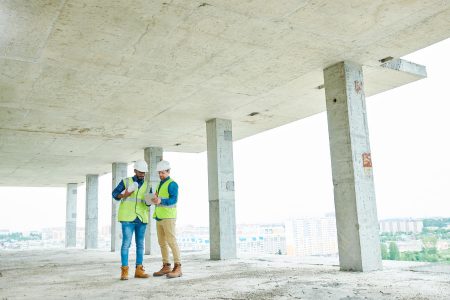 Two men in protective helmets and waistcoats discussing construction plan while standing under ceiling of unfinished building